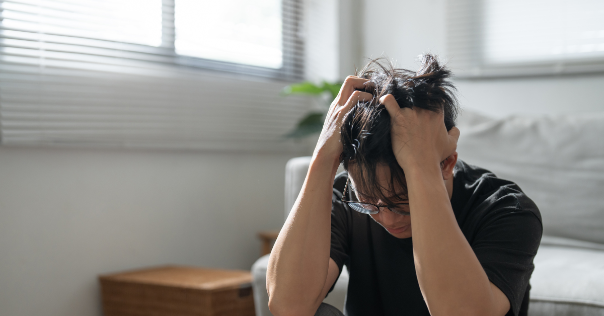 A man sitting indoors with his head down, gripping his hair in distress, conveying a strong sense of stress or anxiety. The soft natural light and neutral surroundings contrast with his emotional state.