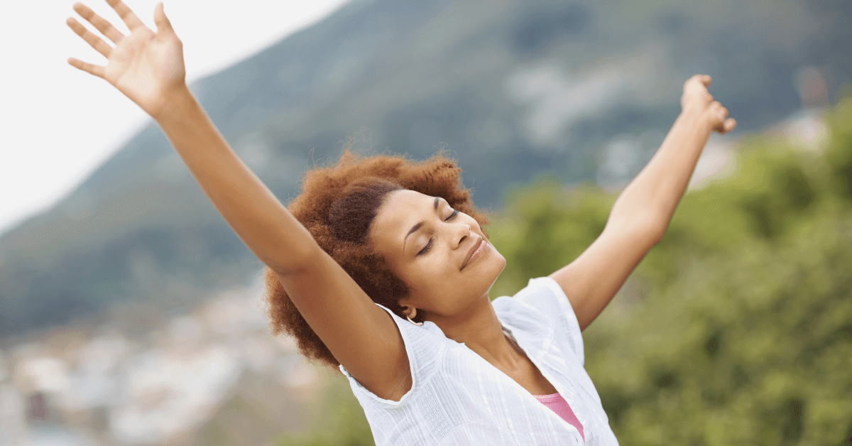 A joyful woman with curly hair and a white blouse stands outdoors with her arms raised, eyes closed, and a serene smile.